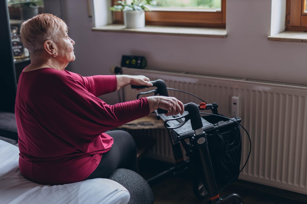 Older woman in a magenta top gripping the handles of a rollator walker beside a window and radiator